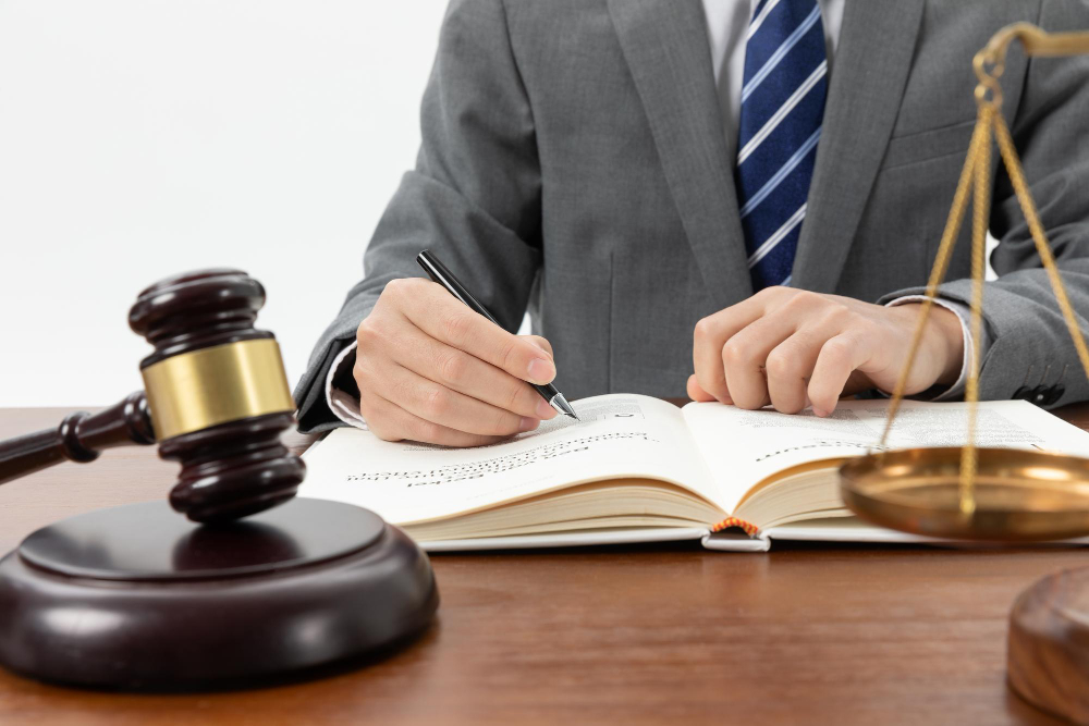 Legal professional in a gray suit writes in an open law book on a desk, with a gavel and scales nearby.