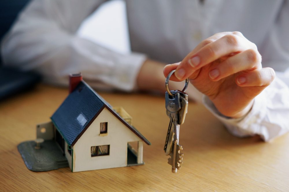 775144 - Gestibensa - Europa News 24h Hand holding a key ring above a miniature house on a wooden table, symbolizing home ownership or real estate.