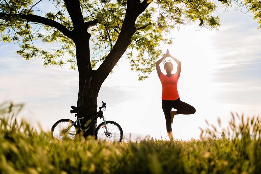 Person in a bright orange top performs a tree pose on one leg beside a bicycle leaning against a tree in a sunny park