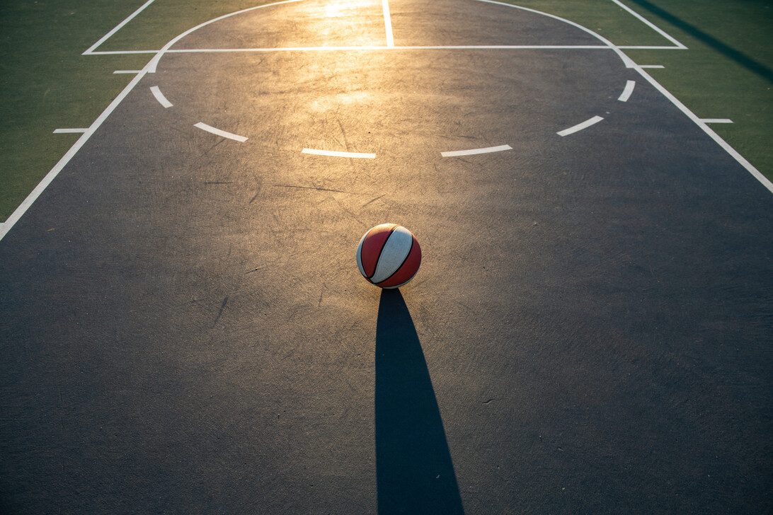 775540 Mtzusa - Europa News 24h Basketball resting on an outdoor court with white lines, low evening light, and a long shadow.