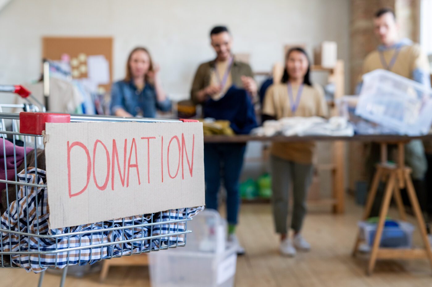 Shopping cart with donation clothes against row of volunteers standing by table - Europa News 24h Cart filled with clothing stools labeled DONATION in a charity drive, with volunteers sorting items in the background.