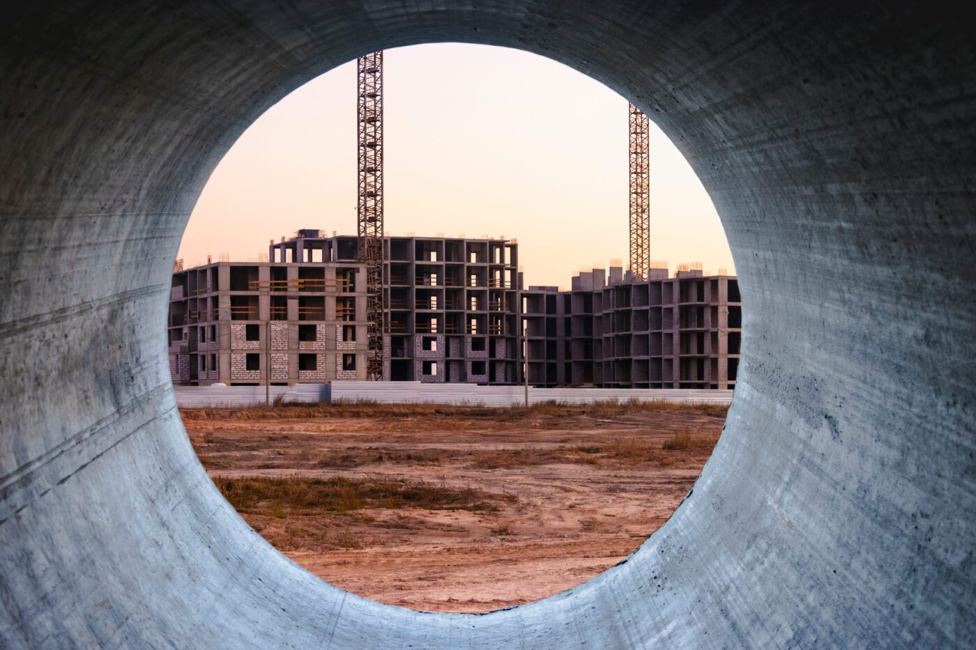 776271 Inside the team - Europa News 24h Construction site framed by a circular concrete pipe, showing unfinished multi-story buildings and two cranes at sunset.