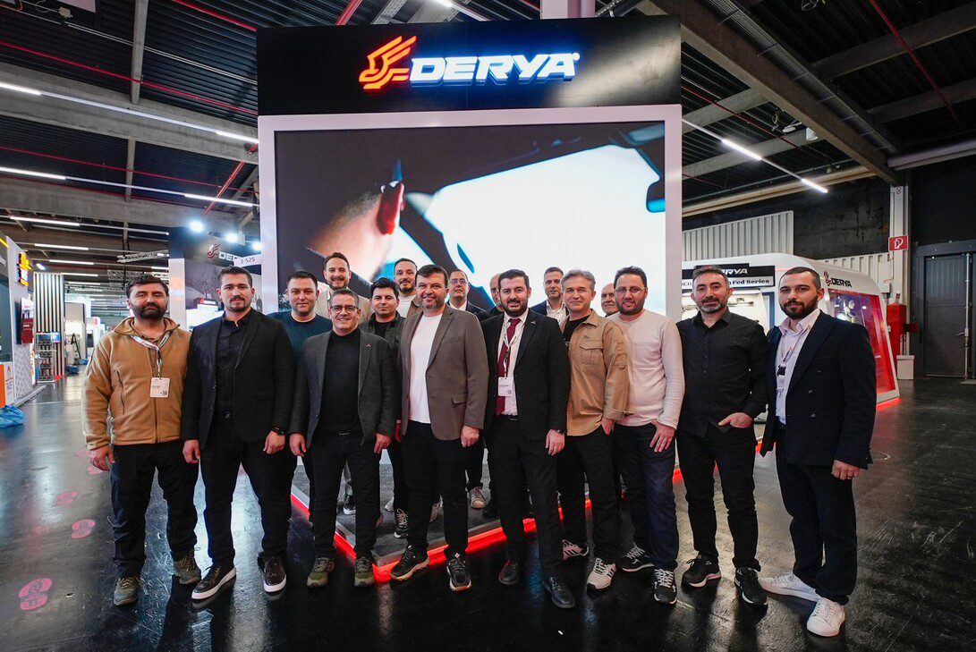 Derya Arms - IP - Europa News 24h Group of men posing in front of a DERYA booth with a large screen at an indoor exhibition hall.