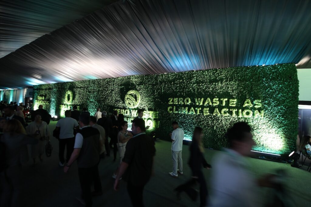 Indoor event with a green hedge wall that reads 'Zero Waste Climate Action' and groups of people mingling in front of it.