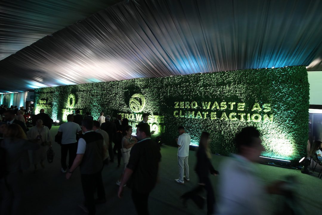 Indoor event with a green hedge wall that reads 'Zero Waste Climate Action' and groups of people mingling in front of it.