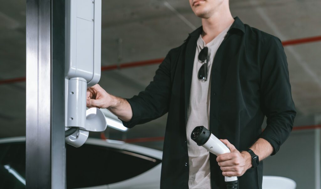 Man connects an electric vehicle charging cable to a wall unit beside a parked car in a garage.