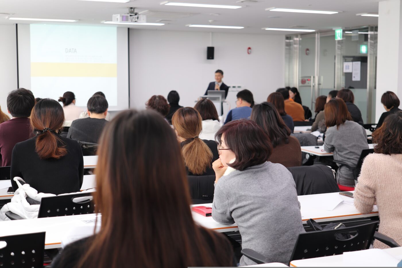 776673 - CFP IN - Europa News 24h Presenter at a podium giving a talk to a full classroom audience of adults seated at desks with a projection screen behind him.
