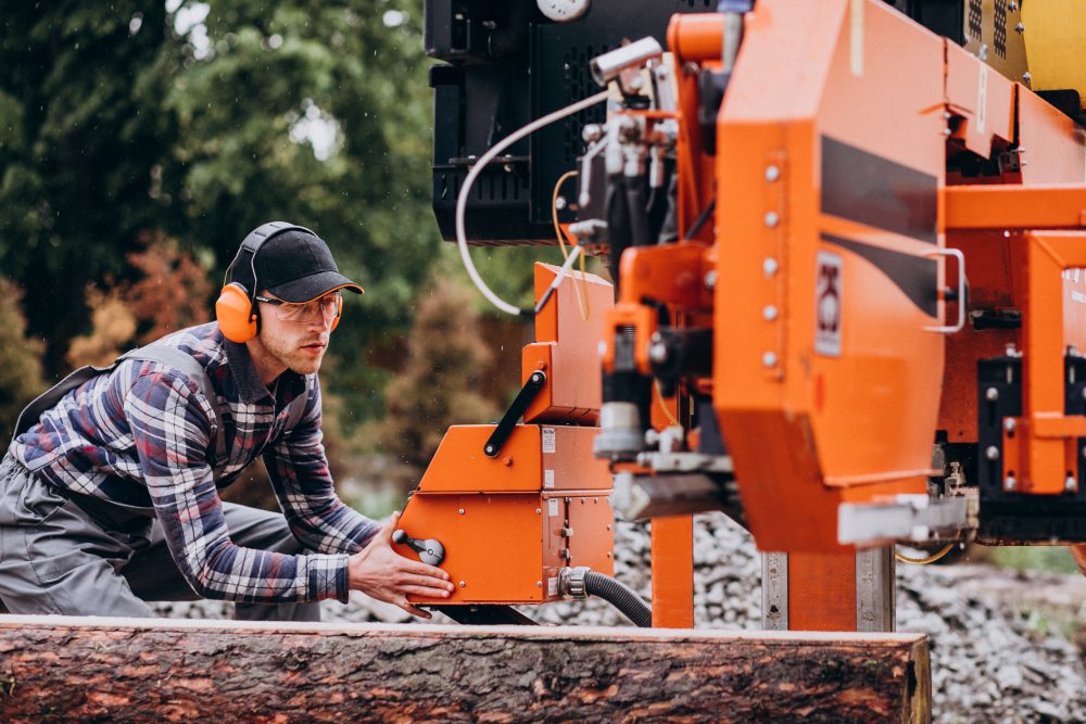 777573 - Pressoil Ibérica SL - Europa News 24h Worker in a plaid shirt and orange ear protection operates a large orange log-cutting machine outdoors, guiding a log into the blade.