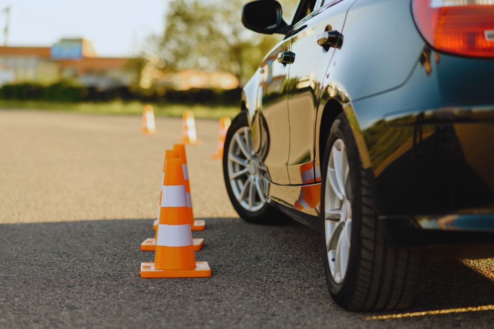 Close-up of a dark car driving around orange and white traffic cones on a parking lot asphalt surface.