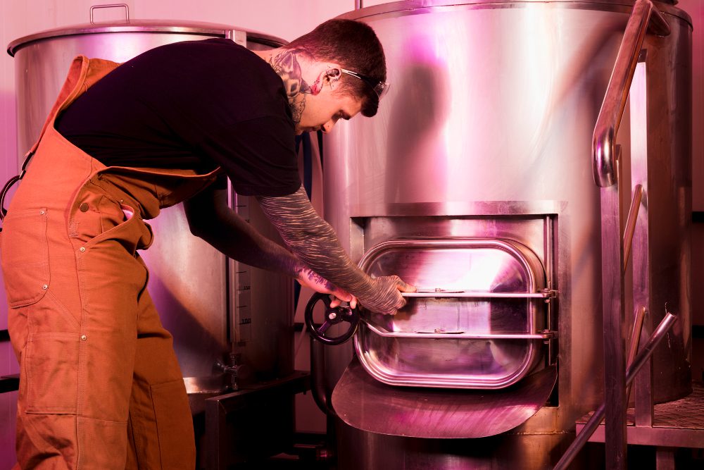 Worker in orange coveralls opens a large stainless steel industrial oven using a hand wheel, in a processing facility