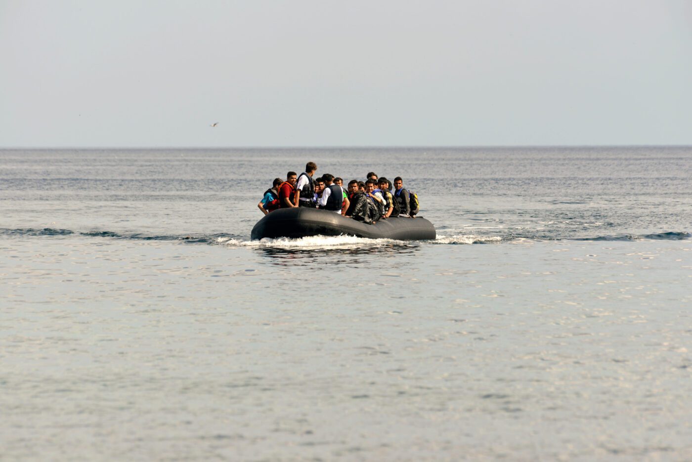 Crowded inflatable boat with about two dozen people in life jackets on calm sea, likely migrants at sea.