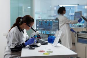 Vivotecnia - IP - Europa News 24h Scientist wearing gloves and safety goggles uses a microscope at a lab bench with a computer monitor nearby; another researcher works in the background.