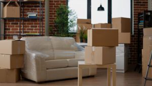 Living room with stacked cardboard boxes and a sofa wrapped in plastic, indicating a move or packing.
