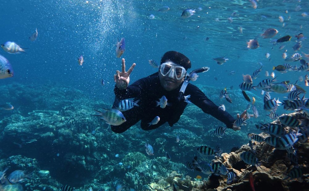 Snorkeler in a wetsuit with mask and snorkel, giving a peace sign while surrounded by tropical fish underwater near a coral reef.