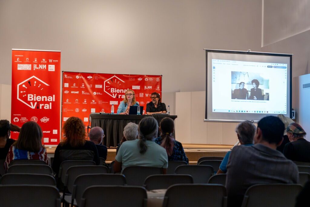 Conference scene with two speakers at a table on stage, red Bienal Viral banners behind them, audience seated, and a projector screen showing a slide.