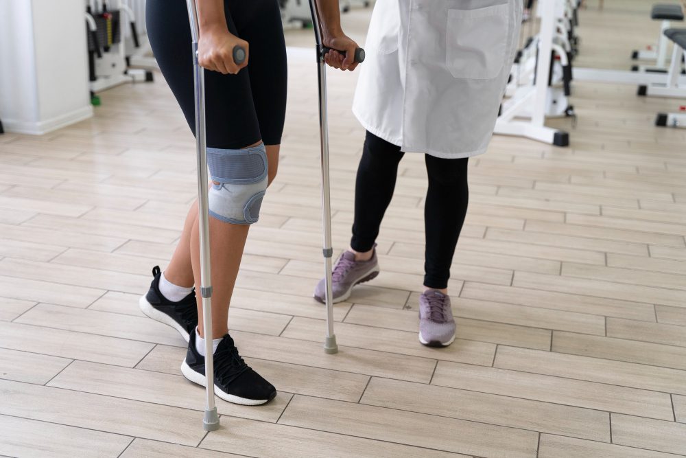 784089 - Ortopedia Neoforma - Europa News 24h Person using forearm crutches with a knee brace, assisted by a clinician in a gym rehabilitation setting.