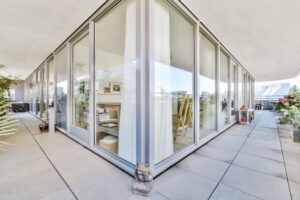Glass-walled living space on a sunlit terrace with tall windows, sheer curtains, and potted plants along the edge.