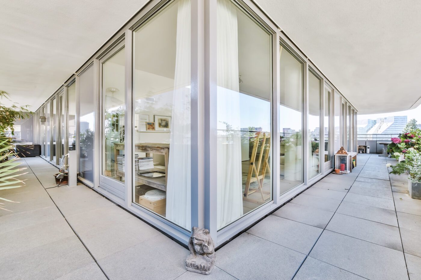 A spacious balcony of a luxury house - Europa News 24h Glass-walled living space on a sunlit terrace with tall windows, sheer curtains, and potted plants along the edge.