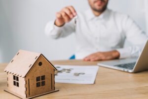 Person in a white shirt hands over a key toward a wooden house model on a desk, with a laptop and papers nearby, illustrating a real estate transaction concept.