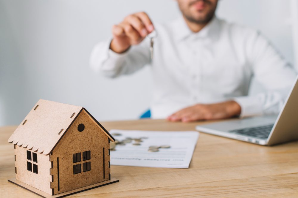 Person in a white shirt hands over a key toward a wooden house model on a desk, with a laptop and papers nearby, illustrating a real estate transaction concept.